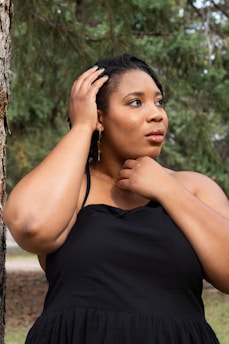 A woman is standing outdoors with her hands gently touching her head and neck, wearing a black sleeveless dress. She appears thoughtful, gazing off into the distance. The background consists of lush green trees, suggesting a natural setting.