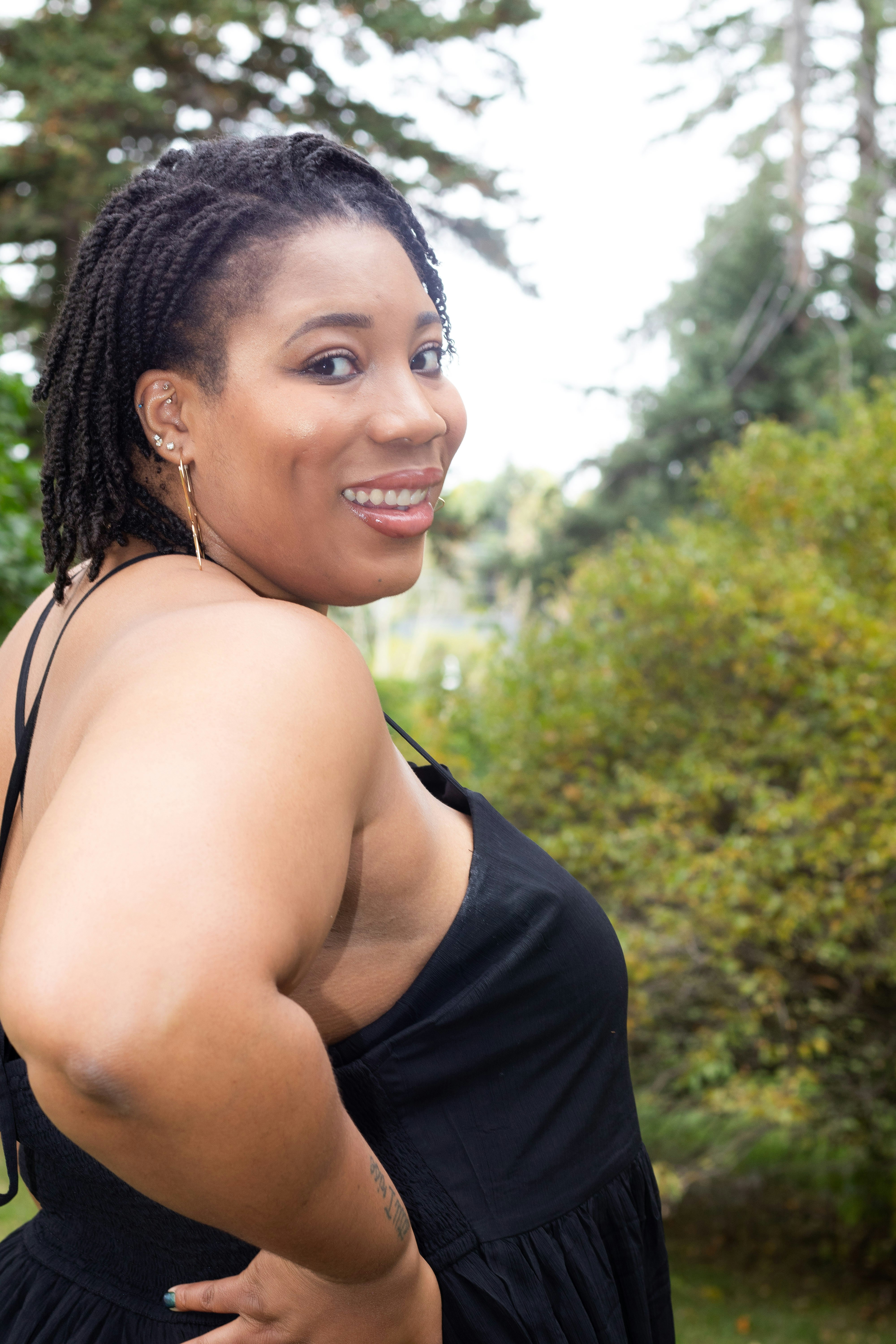 A cheerful woman with a braided hairstyle, wearing a peach blouse, standing outdoors with greenery behind.