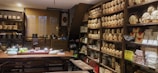 A cozy tea shop interior with shelves lined with neatly stacked tea packages. A large wooden table is set with teaware, teacups, and a bowl of grapes. Various pottery and decorative items are displayed around the room, giving a traditional and inviting atmosphere.