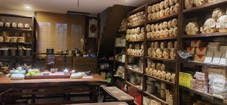 A cozy tea shop interior with shelves lined with neatly stacked tea packages. A large wooden table is set with teaware, teacups, and a bowl of grapes. Various pottery and decorative items are displayed around the room, giving a traditional and inviting atmosphere.