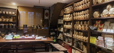 A cozy tea shop interior with shelves lined with neatly stacked tea packages. A large wooden table is set with teaware, teacups, and a bowl of grapes. Various pottery and decorative items are displayed around the room, giving a traditional and inviting atmosphere.