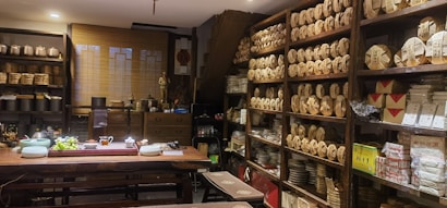 A cozy tea shop interior with shelves lined with neatly stacked tea packages. A large wooden table is set with teaware, teacups, and a bowl of grapes. Various pottery and decorative items are displayed around the room, giving a traditional and inviting atmosphere.
