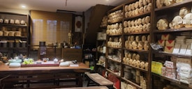 A cozy tea shop interior with shelves lined with neatly stacked tea packages. A large wooden table is set with teaware, teacups, and a bowl of grapes. Various pottery and decorative items are displayed around the room, giving a traditional and inviting atmosphere.