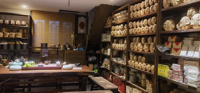 A cozy tea shop interior with shelves lined with neatly stacked tea packages. A large wooden table is set with teaware, teacups, and a bowl of grapes. Various pottery and decorative items are displayed around the room, giving a traditional and inviting atmosphere.