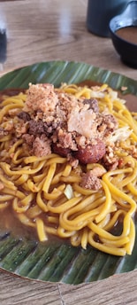 A plate of yellow noodles topped with crispy fried garnish and meat, served on a banana leaf. Accompanied by a bowl in the background on a wooden table.