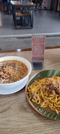 A bowl of noodle soup and a plate of stir-fried noodles are placed on a wooden table. The noodle soup has a thick broth and is garnished with crispy toppings. The stir-fried noodles are served on a banana leaf and also garnished with crispy toppings. There is a metal table number stand in the background displaying the number '03'. The dining area in the background has several tables and chairs.