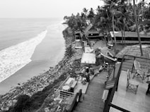 A serene coastline view features a calm ocean meeting a rocky shore. Palm trees and thatched-roof structures line the coast, with a staircase leading down to the water. Patio furniture, including tables and chairs, is visible, suggesting an outdoor dining area. The scene conveys a tranquil, tropical atmosphere.