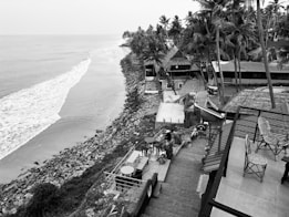 A serene coastline view features a calm ocean meeting a rocky shore. Palm trees and thatched-roof structures line the coast, with a staircase leading down to the water. Patio furniture, including tables and chairs, is visible, suggesting an outdoor dining area. The scene conveys a tranquil, tropical atmosphere.