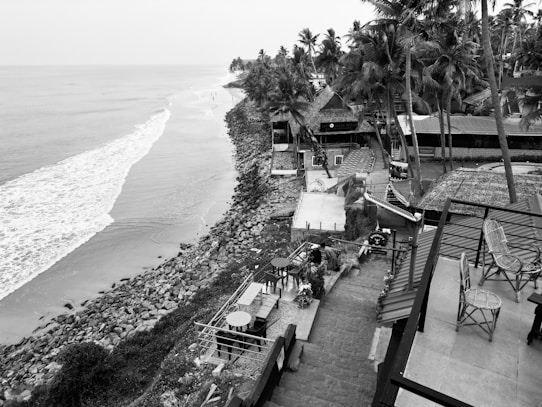 A serene coastline view features a calm ocean meeting a rocky shore. Palm trees and thatched-roof structures line the coast, with a staircase leading down to the water. Patio furniture, including tables and chairs, is visible, suggesting an outdoor dining area. The scene conveys a tranquil, tropical atmosphere.