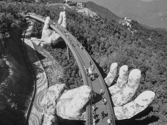 A large pedestrian bridge supported by two giant stone hands is surrounded by a lush mountainous landscape. People are walking along the bridge, enjoying the view. The environment is a blend of natural beauty and artistic architecture.