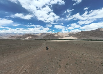 A vast, barren landscape with rugged, rocky mountains in the background under a sky filled with scattered white clouds. In the foreground, a lone person rides a motorcycle along a dusty path cutting through the desolate terrain.