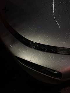 Raindrops are scattered across the surface of a sleek, metallic car hood, reflecting ambient light and creating a glossy appearance. The curvature of the vehicle is highlighted by the light's reflection, while the dark background contrasts with the shiny metallic finish.