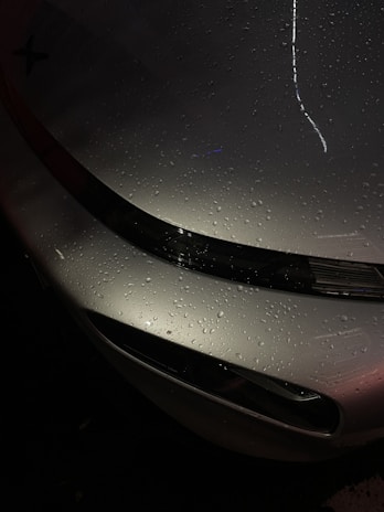 Raindrops are scattered across the surface of a sleek, metallic car hood, reflecting ambient light and creating a glossy appearance. The curvature of the vehicle is highlighted by the light's reflection, while the dark background contrasts with the shiny metallic finish.