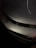 Raindrops are scattered across the surface of a sleek, metallic car hood, reflecting ambient light and creating a glossy appearance. The curvature of the vehicle is highlighted by the light's reflection, while the dark background contrasts with the shiny metallic finish.
