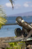A bronze statue of a surfer is positioned close to a body of water. The statue is adorned with plumeria flowers tucked behind the ear, surrounded by green palm leaves. The background consists of the sea and a blurred horizon.