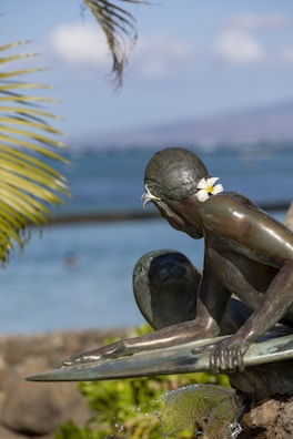 A bronze statue of a surfer is positioned close to a body of water. The statue is adorned with plumeria flowers tucked behind the ear, surrounded by green palm leaves. The background consists of the sea and a blurred horizon.
