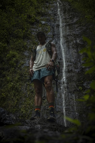 A person stands in front of a tall, narrow waterfall, surrounded by lush greenery. The individual is wearing a backpack, shorts, and hiking boots, and appears to be in a tranquil, natural setting.