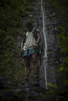A person stands in front of a tall, narrow waterfall, surrounded by lush greenery. The individual is wearing a backpack, shorts, and hiking boots, and appears to be in a tranquil, natural setting.