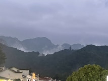 A tranquil landscape featuring rolling hills and mountains shrouded in mist and clouds. In the foreground, traditional-style buildings with tiled roofs are visible. A solitary pagoda stands on a hillside, adding an element of historical or cultural significance.