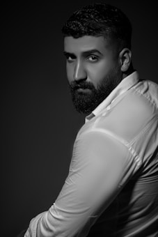 A portrait of a person with a beard, wearing a white shirt, posed against a dark background. The monochrome image highlights expressions and textures.