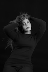 A black and white portrait of a woman posing with her arms raised, fingers running through her long hair. She is wearing a dark long-sleeve top and gazes directly at the camera with an expression of confidence and elegance. The background is plain, emphasizing the subject.