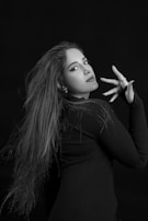 A woman with long hair poses in a stylish manner against a dark background. Her expression is confident, and she's wearing a black top. Her makeup is bold, emphasizing her eyes and lips. One hand is elegantly raised near her face, with fingers slightly splayed.