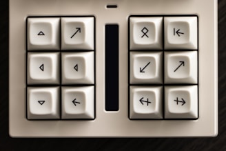A white keypad featuring eight square-shaped keys with unique arrow symbols pointing in various directions. The keys are arranged in a 2x4 grid and surrounded by a black border, with a narrow and elongated rectangular screen or touchpad situated in the center.