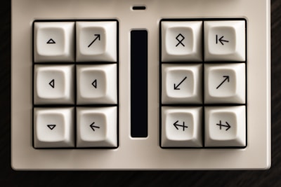 A white keypad featuring eight square-shaped keys with unique arrow symbols pointing in various directions. The keys are arranged in a 2x4 grid and surrounded by a black border, with a narrow and elongated rectangular screen or touchpad situated in the center.