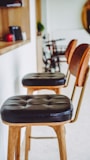 A well-lit space featuring two wooden bar stools with black leather cushions. The background shows a shelf with items, a blurred bicycle, and green plant decor. The bar stools are aligned along a white wall next to a wooden counter.