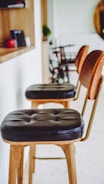 A well-lit space featuring two wooden bar stools with black leather cushions. The background shows a shelf with items, a blurred bicycle, and green plant decor. The bar stools are aligned along a white wall next to a wooden counter.