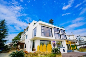 A modern, white two-story building with large glass windows, set against a vibrant blue sky with scattered clouds. The structure incorporates traditional architectural elements, visible in the surrounding buildings and rooflines. Lush greenery, including trees and bushes, surround the building, contributing to a serene and inviting atmosphere.