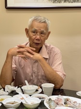 An elderly man with short gray hair sits at a table, wearing a light pink shirt. His hands are clasped together, and he is surrounded by several small dishes and a white cup. The background features a piece of framed art on the wall.