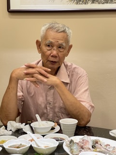 An elderly man with short gray hair sits at a table, wearing a light pink shirt. His hands are clasped together, and he is surrounded by several small dishes and a white cup. The background features a piece of framed art on the wall.