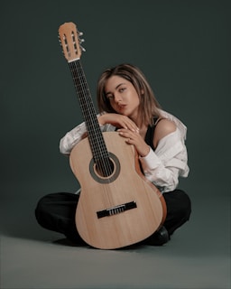 A woman sits on a floor with a classical acoustic guitar positioned vertically in front of her. She is wearing a white off-shoulder shirt and dark pants. The background is a muted gray, and the overall setting has a calm and intimate atmosphere.