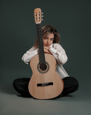 A person is sitting cross-legged on the floor, leaning gently against an acoustic guitar. They are wearing a white shirt and dark pants, with their hair styled in a casual manner. The background is a plain, dark green color, adding a calm and minimalistic touch to the composition.