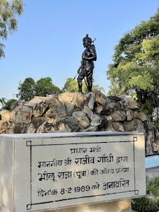 A statue of a man holding a bow and arrow stands atop rocky terrain. The sculpture appears to be made of dark material, possibly metal, and is set against a background of trees and a clear blue sky. At the base of the statue, a plaque is inscribed in Hindi, mentioning a dedication by Rajiv Gandhi dated 8-2-1989.