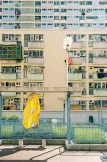 A tall residential building with numerous balconies and windows, some of which have laundry hanging. In the foreground, a bright yellow covering with cartoon illustrations is draped over a chair or small structure. A clean, modern urban atmosphere is present, with a mix of concrete and greenery.