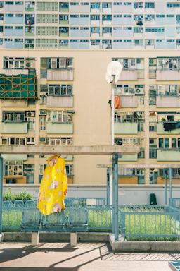 A tall residential building with numerous balconies and windows, some of which have laundry hanging. In the foreground, a bright yellow covering with cartoon illustrations is draped over a chair or small structure. A clean, modern urban atmosphere is present, with a mix of concrete and greenery.