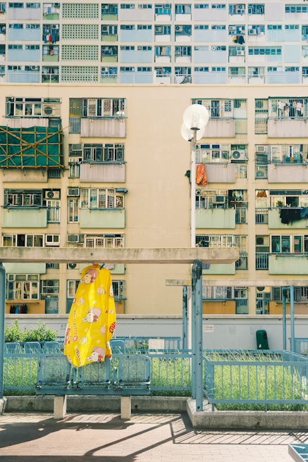A tall residential building with numerous balconies and windows, some of which have laundry hanging. In the foreground, a bright yellow covering with cartoon illustrations is draped over a chair or small structure. A clean, modern urban atmosphere is present, with a mix of concrete and greenery.