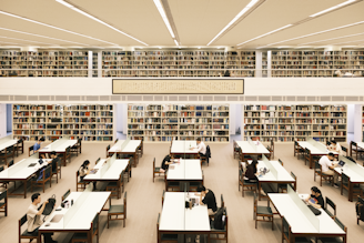 A spacious library interior filled with rows of bookshelves and tables. People are sitting at the tables, reading or working on laptops. The library has a high ceiling with fluorescent lights, and a framed scroll with writing is displayed above.