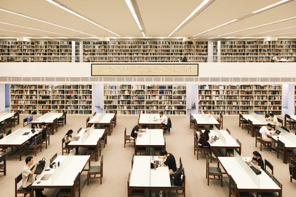 A spacious library interior filled with rows of bookshelves and tables. People are sitting at the tables, reading or working on laptops. The library has a high ceiling with fluorescent lights, and a framed scroll with writing is displayed above.