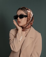 A woman is wearing a headscarf with geometric patterns, oversized black sunglasses, and a beige blazer. Her pose is thoughtful, with one hand placed on her cheek.