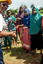 A group of people gathered outdoors, with women and children dressed in colorful clothing and headscarves. They appear to be engaged in a discussion or activity. The setting is grassy with trees in the background, indicating a rural or natural environment. A person in the foreground is partially visible, wearing a yellow cap and speaking animatedly.