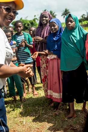 A group of people gathered outdoors, with women and children dressed in colorful clothing and headscarves. They appear to be engaged in a discussion or activity. The setting is grassy with trees in the background, indicating a rural or natural environment. A person in the foreground is partially visible, wearing a yellow cap and speaking animatedly.