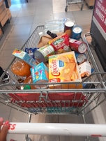 A shopping cart filled with a variety of groceries, including bottled water, canned beverages, a loaf of bread, pasta, orange juice, snacks, and crackers. The cart is situated in what appears to be a supermarket aisle, with tiles on the floor and another trolley visible in the background.
