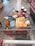 A shopping cart filled with a variety of groceries, including bottled water, canned beverages, a loaf of bread, pasta, orange juice, snacks, and crackers. The cart is situated in what appears to be a supermarket aisle, with tiles on the floor and another trolley visible in the background.