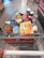 A shopping cart filled with a variety of groceries, including bottled water, canned beverages, a loaf of bread, pasta, orange juice, snacks, and crackers. The cart is situated in what appears to be a supermarket aisle, with tiles on the floor and another trolley visible in the background.