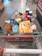 A shopping cart filled with a variety of groceries, including bottled water, canned beverages, a loaf of bread, pasta, orange juice, snacks, and crackers. The cart is situated in what appears to be a supermarket aisle, with tiles on the floor and another trolley visible in the background.