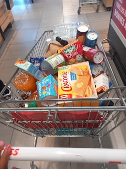 A shopping cart filled with a variety of groceries, including bottled water, canned beverages, a loaf of bread, pasta, orange juice, snacks, and crackers. The cart is situated in what appears to be a supermarket aisle, with tiles on the floor and another trolley visible in the background.