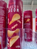 Cylindrical red containers with bold white text displaying 'SAVEUR BACON' and an image of chips and bacon strips are lined up on a shelf. The packaging indicates a 170g size with nutritional information and branding visible.